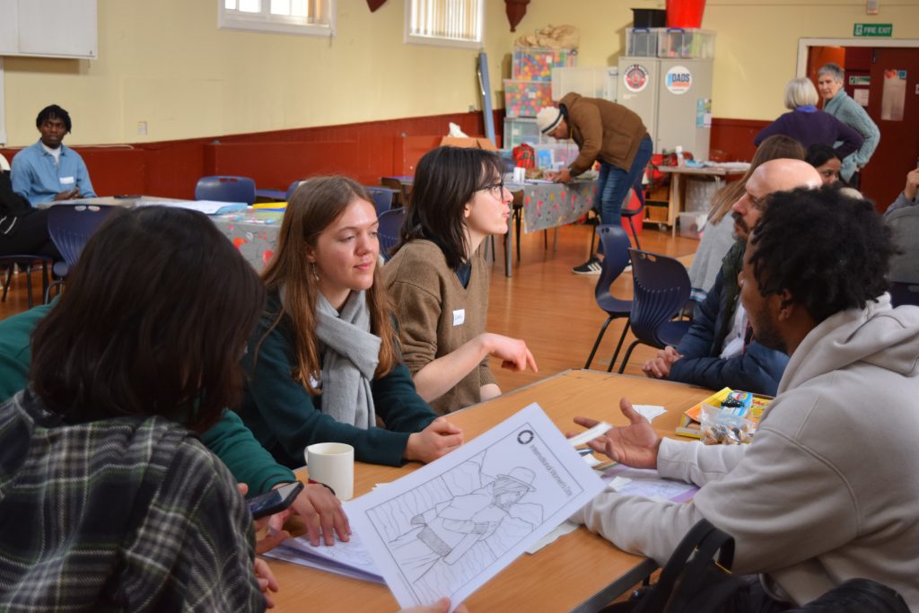Men and women sitting around a table in a busy hall talking and drawing.