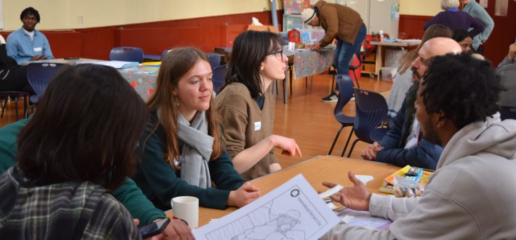 Men and women sitting around a table in a busy hall talking and drawing.