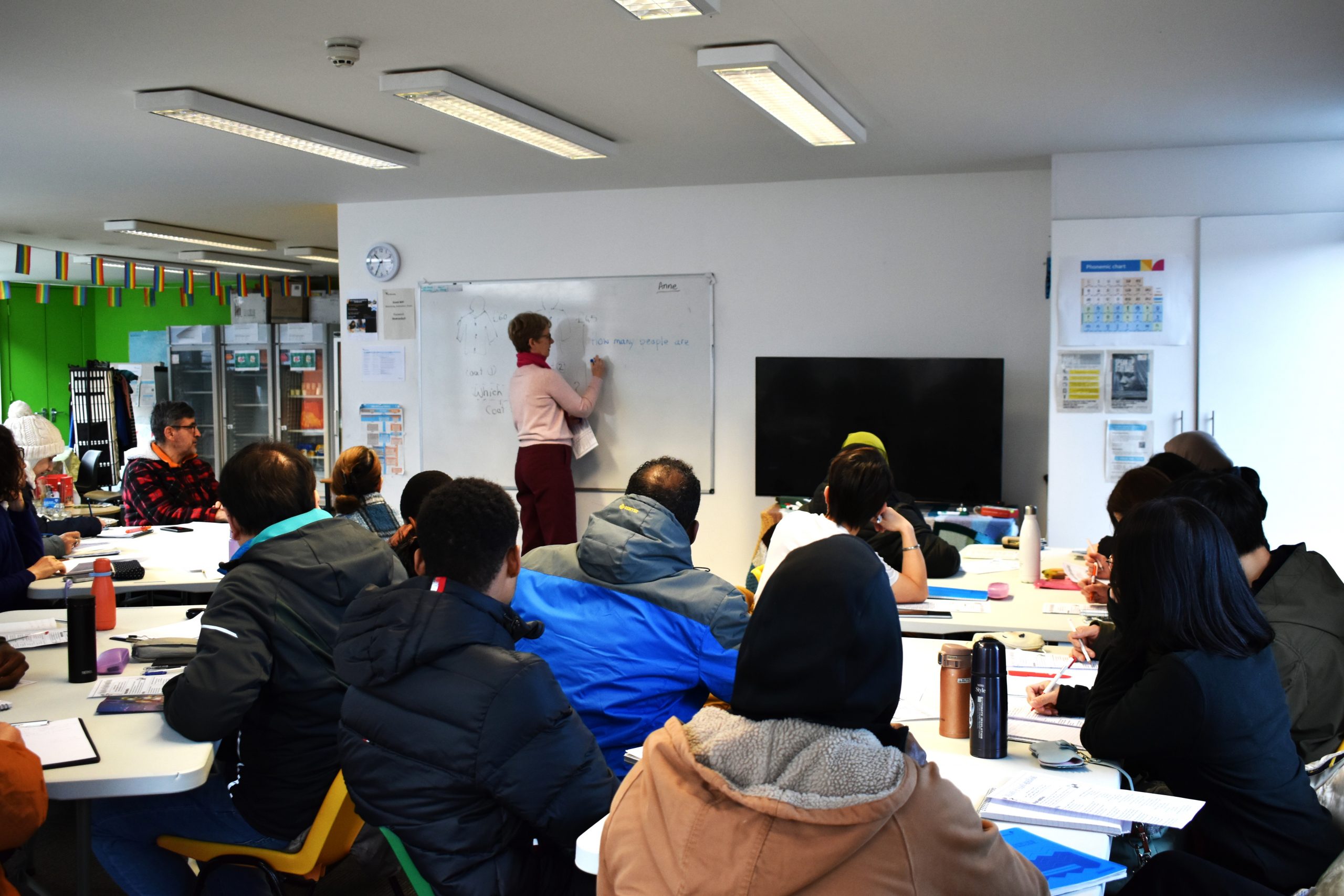 People sitting in the Welcoming classroom learning English looking at a teacher writing on a whiteboard.