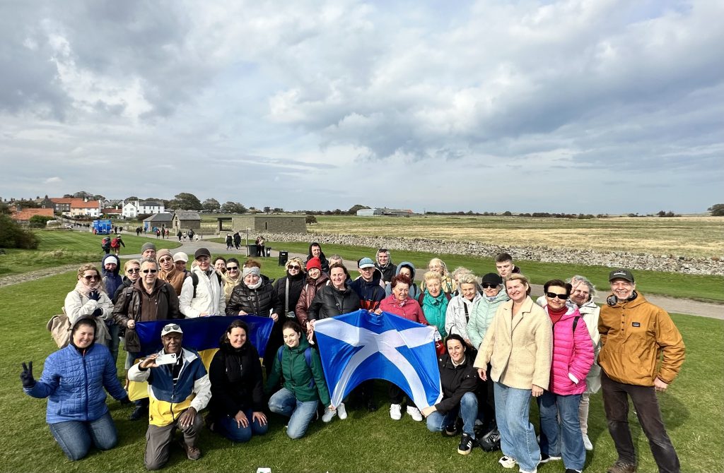 A group of around 40 people gathered outdoors on a grassy field during a trip to Holy Island, Berwick-upon-Tweed. They are smiling at the camera, some holding Ukrainian and Scottish flags. The background shows stone walls, houses, and open countryside under a cloudy sky.