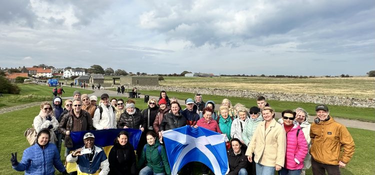 A group of around 40 people gathered outdoors on a grassy field during a trip to Holy Island, Berwick-upon-Tweed. They are smiling at the camera, some holding Ukrainian and Scottish flags. The background shows stone walls, houses, and open countryside under a cloudy sky.
