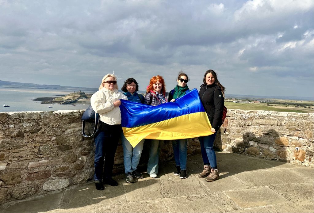 Five women stand together holding a large Ukrainian flag in front of a stone wall at Lindisfarne Castle. Behind them is a wide view of the coastline, sea, and distant landscape under a cloudy sky.