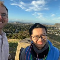 Two men standing on a high hill overlooking Edinburgh facing the camera. They are smiling.